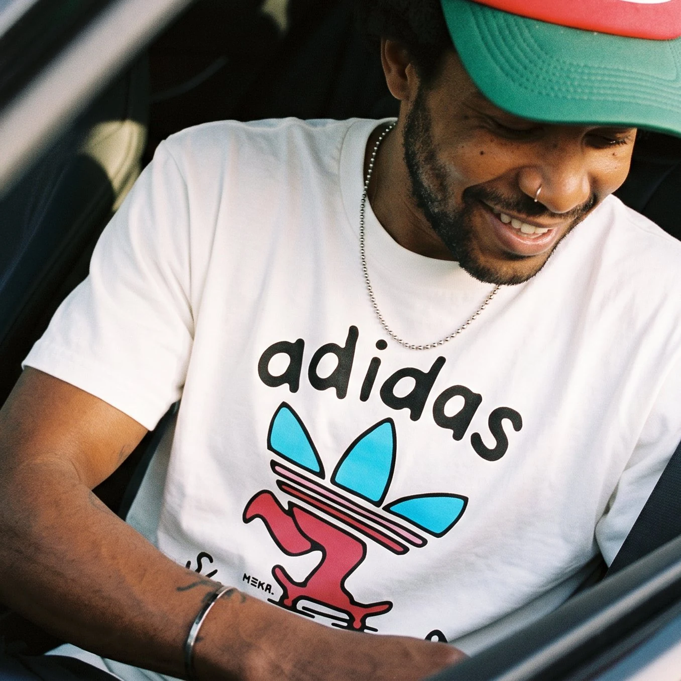 Smiling man in a car wearing the white adidas trefoil tee — the trefoil rebuilt as a Keep Pushing skate character, MEKA. signature
