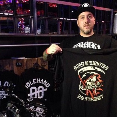 Holding up a Sons of Eighties tee at the shop, bikes behind