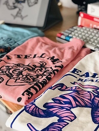 Printed tees stacked on the desk next to the screen
