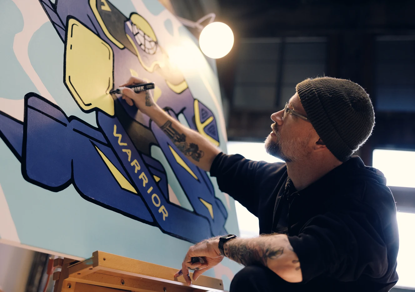 The artist at the easel, brush to the canvas, laying in the goalie's purple pads under a work lamp